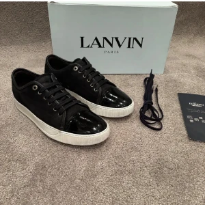 Svarta sneakers från Lanvin med lackdetalj - Snygga svarta sneakers från Lanvin med vit sula och glansig lackad tå. Skorna är UK 7 som motsvara 41-42 i EU. Skorna har klassisk snörning, metallöljetter och en stilren design. Ovandelen är i mocka och detaljer i skinn, vilket ger en lyxig känsla. Perfekta för dig som gillar exklusiva stil