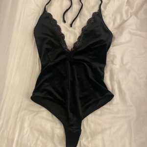 Svart spetsdetaljerad body från BikBok - Supersnygg svart body från BikBok i storlek S. Modellen har djup v-ringning med fina spetsdetaljer längs kanterna och smala knytband i nacken. Materialet är mjukt och stretchigt, vilket ger en bekväm passform och en elegant look.