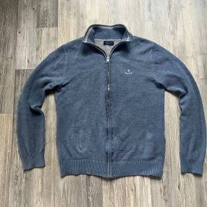 Blå stickad kofta från GANT - Säljer en blå stickad kofta från GANT i storlek S. Koftan har hel dragkedja, ribbade muddar och ståkrage med vita ränder. GANT-logga broderad på bröstet. Perfekt för lager-på-lager och chillare dagar.