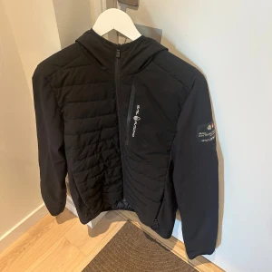Svart pufferjacka från Sail Racing - Svart pufferjacka från Sail Racing med huva och quiltad framsida. Jackan har dragkedja framtill, en bröstficka med dragkedja och logga på bröstet samt ärmen. Perfekt för kyligare dagar. knappt använd 