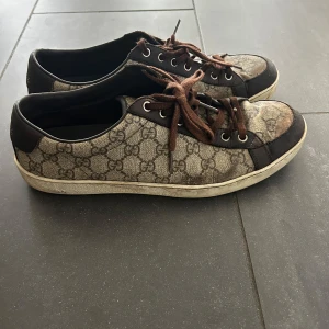 Gucci sneakers med GG-mönster - Snygga sneakers från Gucci med klassiskt GG-mönster i beige och brunt. Skorna har bruna skinn- och textildetaljer, bruna snören och vit sula med unikt mönster undertill. Perfekta för dig som vill ha en lyxig och tidlös look. Stl 10 motsvarar 43/44