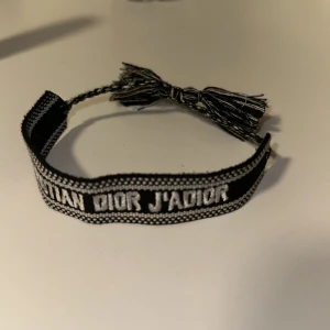 Tyg armband - Snyggt svart och vitt armband med texten Christian Dior broderad. Armbandet har flätade band och två tofsar som detalj, vilket ger en cool och trendig vibe. Perfekt accessoar för att lyfta din outfit.