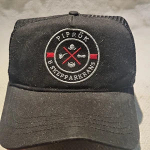 Svart keps från Piprök & Skepparkrans - Svart truckerkeps med broderad logga framtill i vitt, rött och grått. Kepsen har böjd skärm, justerbar snapback och mesh baktill för extra ventilation. Tillverkad i 60% bomull och 40% polyester. Perfekt för en chill och stilren look.