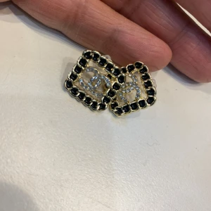  örhängen med svart och guld - Snygga örhängen i fyrkantig form med guldfärgad ram och svarta detaljer. I mitten sitter den klassiska som täckt av glittrande stenar. Perfekta för dig som vill sticka ut med lyxig vibe och ikonisk design.