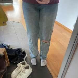 Ljusblå jeansbyxor från Monki - Säljer ett par ljusblå jeansbyxor från Monki med klassisk femficksdesign och raka ben. Byxorna har en tydlig slitning med hål på vänster knä och är tillverkade i bomull. Perfekta för en avslappnad och trendig look.