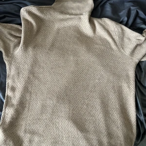 Beige stickad tröja med struktur - Säljer en beige stickad tröja med snyggt strukturerat mönster och ribbade muddar. Tröjan har hög krage och en liten svart detalj vid nederkanten. Perfekt för lager-på-lager under kyliga dagar.