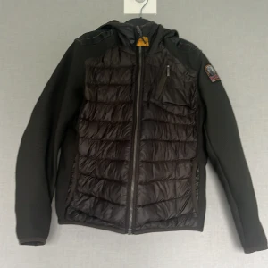 Svart hybridjacka från Parajumpers - Snygg svart hybridjacka från Parajumpers med quiltad front och rygg, huva och coolt PJS-märke på ärmen. Jackan har dragkedja framtill, bröstficka med dragkedja och sportig känsla. Perfekt för kyliga dagar när du vill ha både stil och funktion.