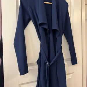 Marinblå långklänning med bälte - Snygg marinblå långklänning med långa ärmar och ett brett, draperat slag framtill. Klänningen har ett matchande bälte i midjan som knyts för en markerad siluett. Perfekt för dig som gillar stilrena och enkla plagg med en twist.