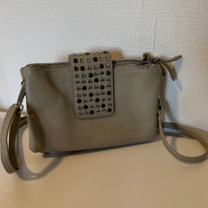 Beige axelväska med nitar och dragkedja - Snygg beige axelväska i skinnimitation med silverfärgade nitar på locket. Väskan har två stora fack med dragkedja och en justerbar axelrem. Perfekt storlek för mobil, plånbok och småprylar. Enkel men cool design som sticker ut. Helt ny, aldrig använt. 