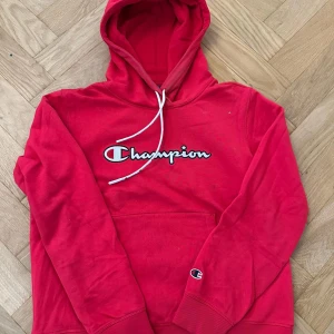 Röd Champion hoodie med huva - Röd hoodie från Champion med stor logga broderad på bröstet och klassisk känguruficka. Tröjan har huva med vit snörning och ribbade muddar. Perfekt för chill dagar eller streetwear-stil.