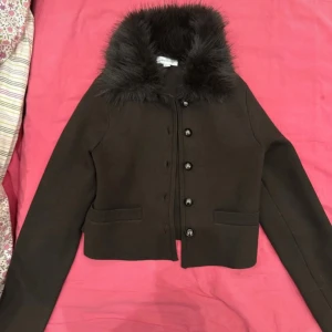 Brun cardigan jacka med fuskpäls - Mörkbrun cardigan jacka från New Edition med fluffig fuskpäls runt kragen. Jackan är croppad, har långa ärmar, två fickor framtill och knäpps med knappar. Perfekt för dig som vill ha en mysig och trendig look.s eller xs