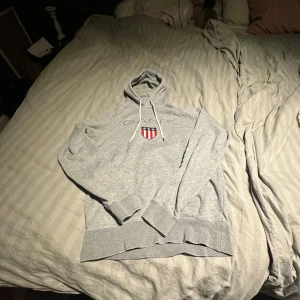Grå hoodie från GANT med brodyr - Säljer en grå hoodie från GANT med broderad logga och sköld på bröstet. Tröjan har snörning i huvan, ribbade muddar och klassisk passform. Perfekt för dig som gillar stilren och sportig look.