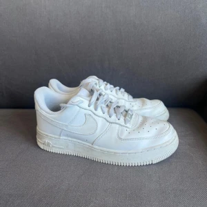 Nike Air Force 1 vita sneakers - Klassiska Nike Air Force 1 sneakers i helvitt skinn med rund tå och snörning. Modellen har den ikoniska swoosh-loggan på sidan och AIR-detalj på sulan. Perfekta för dig som gillar en clean och tidlös look.