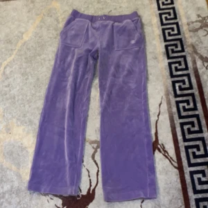 Lila mjukisbyxor från Juicy Couture - Lila mjukisbyxor från Juicy Couture i mjuk sammet med bred resår i midjan, stora fickor fram och en bakficka med knappar. Byxorna har broderad JC-logga på ena fickan och raka ben. Perfekta för en chill och trendig look.