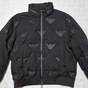 Svart pufferjacka från Armani - Svart pufferjacka från Armani med ikoniska loggor tryckta över hela jackan. Jackan har hög krage, dragkedja framtill och två stora fickor. Perfekt för kalla dagar när du vill ha en stilren och modern look.