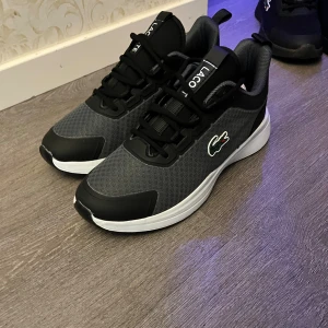 Svarta och grå sneakers från Lacoste - Snygga sneakers från Lacoste i svart och grå mesh med vita sulor och klassisk krokodil-logga på sidan. Skorna har snörning och sportig design, perfekt för en stilren och modern look. Ovandelen är i syntetmaterial och mesh för bra andningsförmåga.