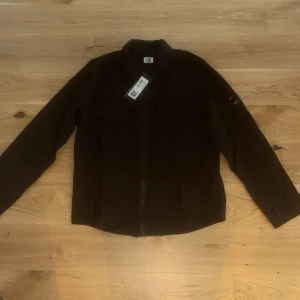 Svart overshirt från C.P. Company - Svart overshirt från C.P. Company i syntetmaterial med klassisk krage, två bröstfickor med knappar och ärmficka med dragkedja och ikonisk linsdetalj. Rak passform och hel dragkedja framtill. Perfekt till höst, vår samt tidig vintertid. Jackan passar dem som har storlek M också då modellen inte är så stor i storlek.