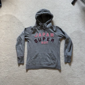 Vintage Japan Superdry hoodie🇯🇵 - Vintage hoodie från Superdry med rött och rosa tryck på bröstet där det står 'JAPAN SUPERDRY'. Tröjan har huva med snörning och en stor magficka. Perfekt för dig som gillar en avslappnad och sportig stil. Storlek M men passar S.