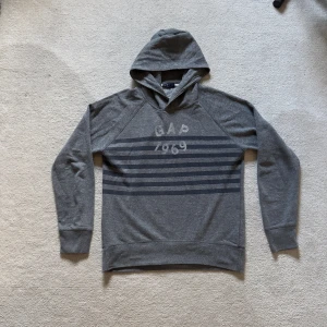 Vintage GAP hoodie med ränder🌊 - Snygg mörkgrå vintage hoodie från GAP med tryck 'GAP 1969' och horisontella svarta ränder på bröstet. Tröjan har huva, långärmad design och ribbade muddar. Perfekt för chill dagar och enkel att matcha med jeans eller joggers.