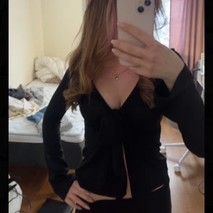 Svart topp med knyt framtill - Säljer en svart topp med djup v-ringning och knyt framtill. Toppen har långa ärmar som är lite vidare ut till. Perfekt för dig som gillar stilrena plagg.