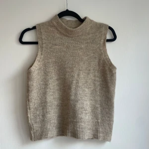 Beige stickad väst från Design by Si - Beige stickad väst med hög krage från Design by Si.  ärmlös och har ribbade detaljer vid hals och nederkant. 