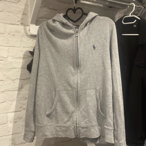 Grå zip hoodie från Polo Ralph Lauren - Grå zip hoodie från Polo Ralph Lauren med dragkedja och huva. Tröjan har två fickor framtill och klassisk blå Polo-logga på bröstet. Tillverkad i mjuk bomull, perfekt för chill dagar. Långärmad och med storlek M. 