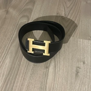 Svart H belte skärp med guldspänne - Säljer ett svart H belte bälte i skinn med ikoniskt H-spänne i guld. Bältet har vit kontrastsöm längs kanterna och en stilren, klassisk design som lyfter vilken outfit som helst. Perfekt för dig som gillar exklusiva accessoarer och vill sticka ut med ett statement piece.