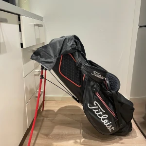Svart golfbag från Titleist med röda detaljer - Säljer en snygg svart golfbag från Titleist med röda och vita detaljer. Väskan har flera fack med dragkedja, praktiska bärremmar och ett regnskydd. Materialet är slitstark syntet och designen har rutmönstrade paneler. Perfekt för dig som vill ha stil på golfbanan.