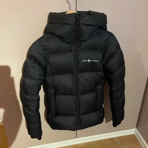 Sail Racing jacka - Säljer en svart dunjacka från Sail Racing med huva och dragkedja framtill. Jackan har en puffig design, logga på bröstet och reflekterande detalj på huvan. Perfekt för kalla dagar och riktigt snygg streetstil.