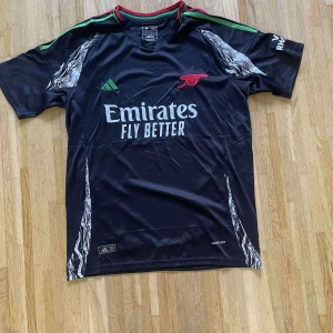 Arsenal svart bortatröja Adidas - Svart Arsenal bortatröja från Adidas med gröna och röda detaljer på krage och axlar. Tryckta loggor, Emirates Fly Better sponsortryck och klubbmärke i rött. Unika vita mönster längs sidorna. Tillverkad i lätt funktionsmaterial.