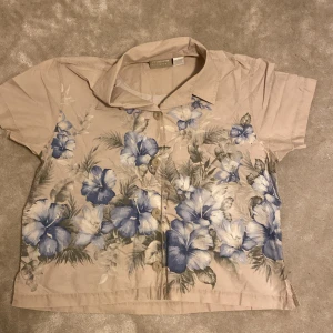 Beige blommig skjorta från Liz Claiborne - Kortärmad skjorta från Liz Claiborne i beige med stora blå och grå blommor. Gjord i 100% bomull. Kort och bred, boxy. Är öppen för ett lägre pris vid snabb affär. Om ni har några frågor skriv gärna!