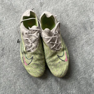 Nike Phantom GX fotbollsskor strl 42 - Nike Phantom GX fotbollsskor i grått och ljusgrönt med svarta och rosa detaljer. Ovandel i stickad syntet med spiralformade mönster, snörning och rund tå. Yttersula med fasta dobbar och rosa Swoosh-logga undertill. Perfekta för dig som vill sticka ut på planen.väldigt använde säljer till ett bökigt pris 