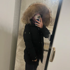 Svart dunjacka med pälsluva - Säljer en svart dunjacka med stor fluffig päls på luvan. Jackan har dragkedja och knappar framtill, samt fickor med dragkedja på sidorna. Lång modell som håller dig varm och har en clean, stilren look. Perfekt för kalla dagar och snygg till vinteroutfiten.