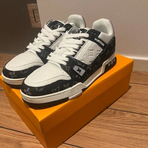 Louis Vuitton svartvita sneakers - Säljer ett par Louis Vuitton sneakers i svart och vitt med klassiskt monogrammönster. Skorna har snörning, platt sula och rund tå. Materialet är en mix av skinn och textil, med tydliga LV-detaljer på sidan och hälen. Perfekt för dig som vill sticka ut med lyxig streetstyle.