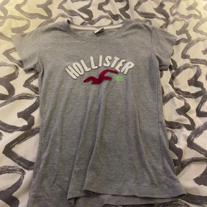Grå Hollister t-shirt med logga - Snygg grå t-shirt från Hollister med broderad vit och röd logga framtill. Klassisk rund halsringning och korta ärmar. Mjuk och skön bomullskvalitet, perfekt för en avslappnad stil.