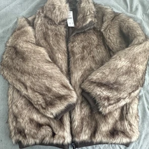 Brun pälsjacka från Urban Outfitters - Säljer en brun pälsjacka från Selected Femme i storlek L. Jackan har mjukt fuskpälsmaterial, ribbade muddar och dragkedja framtill. Perfekt för dig som vill sticka ut med en cozy och trendig look under kyliga dagar. Helt ny!