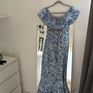 Blommig blå långklänning offshoulder - Säljer en superfin långklänning med blått blommigt mönster och bred volang upptill. Klänningen har offshoulder-design och är perfekt för varma dagar. Materialet känns lätt och luftigt, och den har en smockad midja för skön passform.