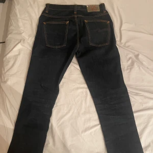 Mörkblå selvedge jeans från Nudie Jeans - Säljer ett par mörkblå jeans från Nudie Jeans med klassisk femficksdesign och kontrastsömmar i orange. Jeansen har rak passform och är tillverkade i kraftigt denimtyg av bomull. Snygga detaljer på bakfickorna och Nudie-logga på bakre läderpatch.