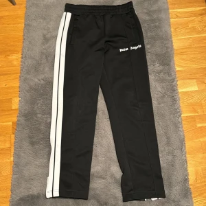 Palm Angels svarta trackpants med vita ränder - Svarta trackpants från Palm Angels med vita dubbla ränder längs sidorna och logga på vänster höft. Byxorna har elastisk midja, raka ben och dragkedja vid benslut. Perfekta för en sportig och trendig look.
