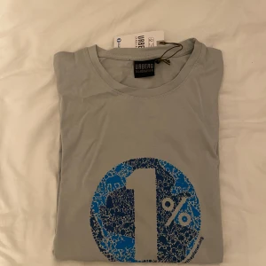 Grå t-shirt med blå print från Urberg - Snygg grå t-shirt från Urberg Skandinavien i storlek M. Framsidan har ett stort blått print med siffran 1% i en cirkel. Klassisk rund hals och korta ärmar. Materialet är mjuk bomull som känns skönt mot huden. Perfekt för dig som gillar enkel och stilren design.