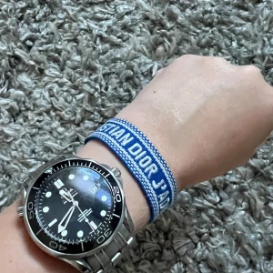 Christian Dior blå armband med tofsar - Säljer ett snyggt blått armband från Christian Dior med texten 'CHRISTIAN DIOR J'ADIOR' broderat i vitt. Armbandet har knytning med två blå och vita tofsar i ändarna och är gjort i ett mjukt tygmaterial. Perfekt för dig som gillar designeraccessoarer och vill sticka ut.
