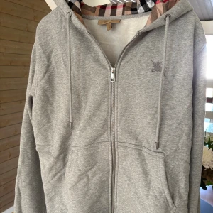 Grå hoodie från Burberry med dragkedja - Grå hoodie från Burberry med klassiskt rutigt foder i huvan, broderad logga på bröstet och dragkedja framtill. Mjuk insida och snörning i huvan. Perfekt för dig som gillar stilrena och lyxiga detaljer.