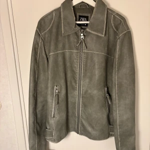 Grå skinnjacka från Zara, storlek L - Säljer en grå skinnjacka från Zara i storlek L. Jackan har klassisk krage, dragkedja framtill och två fickor med dragkedja. Snygga spänndetaljer vid nederkant och ärmar ger extra edge. Perfekt för dig som gillar stilren och cool look.