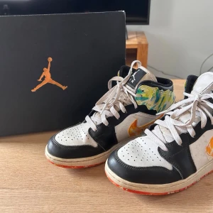 Nike Air Jordan 1 med färgstänk - Unika Nike Air Jordan 1 sneakers med svart och vit ovandel, färgglada detaljer vid ankeln och orangefärgad sula med coola färgstänk. Klassisk snörning och rund tå. Sparsamt använda. Nypris 1700 Kronor. 