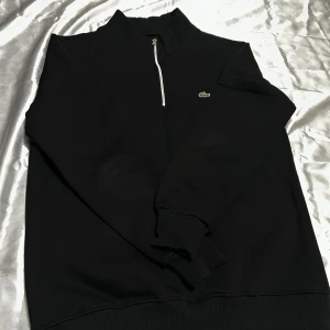 Svart half zip sweatshirt från Lacoste - Svart sweatshirt från Lacoste med half zip och klassisk krokodillogo på bröstet. Tröjan har långärmad design, ribbade muddar och hög krage. Perfekt för dig som gillar stilren och enkel look med en sportig touch.
