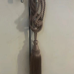 Tazzle - Säljer ett brunt bälte med snurrad design och en stor dekorativ tofs i änden. Bältet är gjort av syntetmaterial och har en unik, bohemisk look som sticker ut. Perfekt för dig som vill ha något annorlunda i ditt hem.