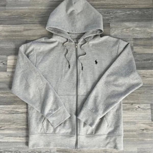 Grå hoodie från Polo Ralph Lauren (Kvitto finns) - Snygg grå hoodie från Polo Ralph Lauren med dragkedja framtill, huva med snörning och klassisk logga broderad på bröstet. Tillverkad i mjuk bomull och har långa ärmar samt praktiska fickor framtill. Perfekt för en chill och avslappnad stil. (kvitto finns)