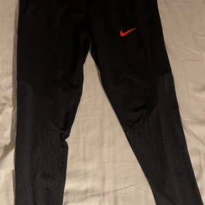 Svarta träningsbyxor från Nike, STL M - Svarta träningsbyxor från Nike med röd swoosh-logga på låret. Byxorna har elastisk midja och paneler med diskret mönster på nedre delen av benen. Tillverkade i ett lätt och syntetiskt material som passar perfekt för sport och träning. 