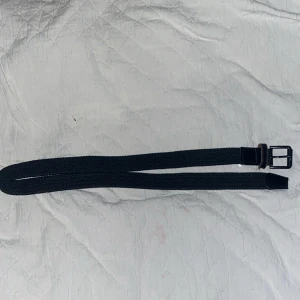 Svart flätat bälte med skinnspänne - Snyggt svart bälte med flätad design och ett stilrent spänne i svart metall. Spännet och ändarna är förstärkta med svart skinn för extra hållbarhet och en clean look. Perfekt för att ge outfiten en enkel men cool touch. 110cm