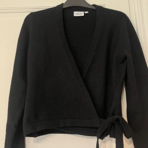 Svart omlottkofta från Kappahl - Svart stickad omlottkofta från Kappahl med knytband i midjan och v-ringad hals. Koftan har lång ärm och är perfekt för lager-på-lager. Enkel och stilren design som funkar till allt. Använd max 6 ggr. Bra skick men bara smått nopprig! Syns knappast